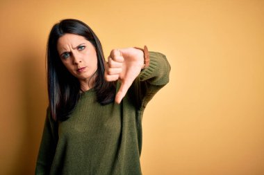 Sarı arka planda yeşil süveter giyen, mavi gözlü genç esmer kadın mutsuz ve kızgın görünüyor, reddediliyor ve baş parmakları aşağıda hareket ediyor. Kötü ifade.