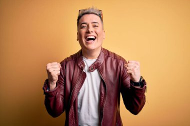 Sarı arka plan üzerinde moda deri ceket ve güneş gözlüğü takan genç, yakışıklı ve modern bir adam kollarını kaldırarak ve açık gözlerle başarıyı kutluyor ve şaşırıyor. Kazanan konsept.