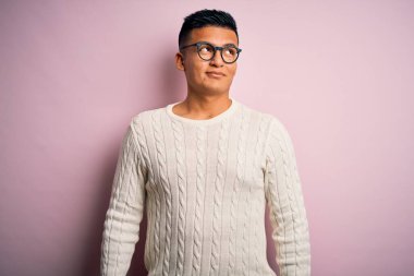 Beyaz süveter giyen, pembe arka planda gözlük takan yakışıklı Latin bir adam gülümsüyor ve başka tarafa bakıyor..