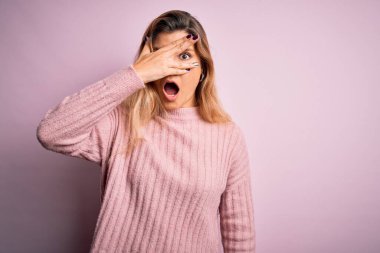 Genç, güzel, sarışın bir kadın, izole edilmiş arka planda pembe bir süveter giyiyor. Şoka giriyor, yüzünü ve gözlerini eliyle kapatıyor, utanmış bir ifadeyle parmaklarından bakıyor..