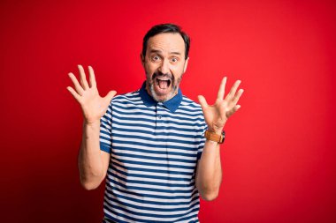 Orta yaşlı, beyaz, sıradan çizgili polo giyen bir adam izole edilmiş kırmızı arka planda durmuş çılgın ve başarıyı kutlarken kolları havada ve gözleri açık bir şekilde heyecanla haykırıyor. Kazanan konsept