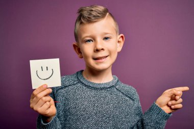 Küçük beyaz bir çocuk kağıt bir notta gülen surat gösteriyor. Mutlu bir mesaj. El ve parmak ile işaret ederek çok mutlu.