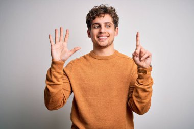 Sarışın, kıvırcık saçlı, beyaz arka planda sıradan bir süveter giyen ve altı numaralı parmaklarını göstererek gülümseyen, kendinden emin ve mutlu genç bir adam..