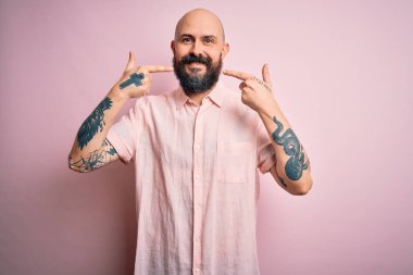 Sakallı ve dövmeli yakışıklı, pembe arka planda günlük gömlekler giyen, neşeli gülümseyen ve parmaklarını dişleriyle ve ağzıyla işaret eden bir adam. Diş sağlığı konsepti.