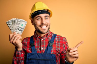 Güvenlik kaskı takan genç müteahhit sarı arkaplan üzerinde dolar tutarken çok mutlu bir şekilde elini ve parmağını kenarı işaret ediyor.