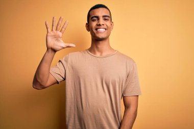 Genç, yakışıklı, Afro-Amerikalı bir adam sarı arka planda dikilen günlük tişörtler giyiyor ve gülümseyen, kendinden emin ve mutlu beş numarayı işaret ediyor..