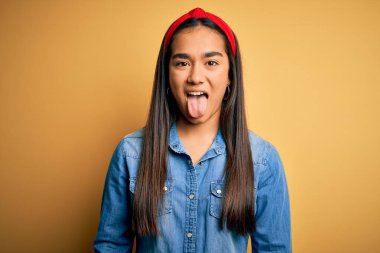 Genç, güzel Asyalı kadın sıradan bir kot gömlek giyiyor ve sarı arka plan üzerine diyabet takmış komik bir ifadeyle dilini dışarı çıkarıyor. Duygu kavramı.