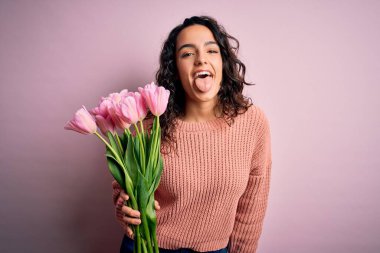 Kıvırcık saçlı, genç ve romantik bir kadın elinde pembe lale buketi, komik bir ifadeyle dilini dışarı çıkartıyor. Duygu kavramı.