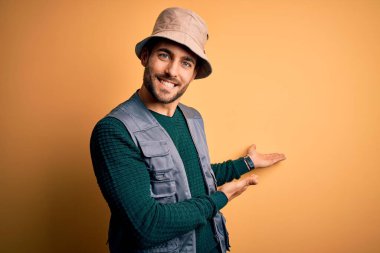 Tatilde sakallı yakışıklı turist sarı arka planda kaşif şapkası takıyor ve gülümseyerek içeri girmeye davet ediyor.