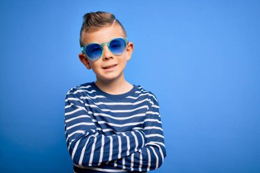 Mavi gözlü, mavi arka planda güneş gözlüğü takan, kollarını kavuşturup kameraya gülümseyen küçük beyaz bir çocuk. Pozitif kişi.