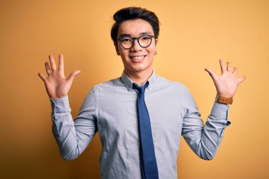 Genç yakışıklı Çinli iş adamı gözlük takıyor ve sarı arka plan üzerine kravat takıyor. 10 numaralı parmaklarıyla işaret ediyor ve gülümsüyor. Kendinden emin ve mutlu..