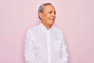 Üst düzey yakışıklı, gri saçlı, zarif tişörtlü, soyutlanmış pembe arka planda, yüzünde gülümseme ve doğal bir ifadeyle bir yana bakan adam. Kendinden emin gülen.