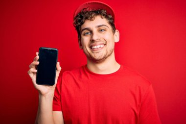 Kıvırcık saçlı, şapkalı, kırmızı arka planda akıllı telefon tutan, mutlu yüzlü, kendine güvenen, gülümseyen ve dişlerini gösteren genç bir teslimatçı.