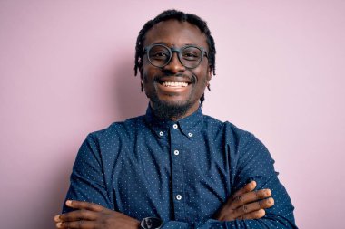 Genç, yakışıklı, Afro-Amerikan bir adam pembe arka planda gözlük takıyor ve kollarını kavuşturup kameraya bakıyor. Pozitif kişi.