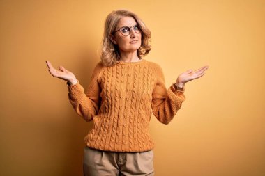 Sarışın, orta yaşlı bir kadın sarı arka planda gözlük takıyor ve kolları ve elleri havada, kafası karışmış bir ifade takınıyor. Şüphe kavramı.