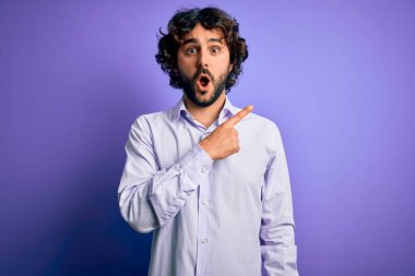 Mor arka plan üzerinde duran sakallı, genç ve yakışıklı bir iş adamı. Parmakla göstererek şaşırmış, ağzı açık, şaşkın bir ifade..