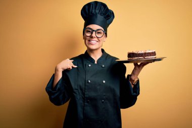 Genç, güzel, esmer fırıncı kadın fırın üniforması giyiyor ve şapkası elinde pastayla kendini gösteren sürpriz bir yüz ifadesi var.