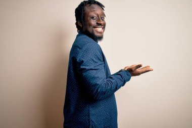 Beyaz arka plan üzerinde duran rahat gömlekli genç, Afro-Amerikan bir adam. Elleri açık, fotokopi alanını gösteren avuç içleri açık, gülümseyerek mutlu bir şekilde gülümseyen bir reklam sunuyor.