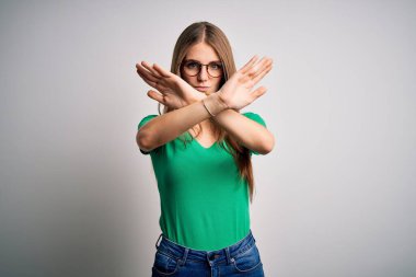 Genç, güzel, kızıl saçlı, yeşil tişörtlü ve gözlüklü bir kadın beyaz arka planda reddedilmiş, kollarını kavuşturmuş, avuç içleri negatif işaretlerle dolu, kızgın yüzlü.