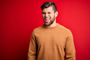 Sakallı ve mavi gözlü genç bir adam kırmızı arka planda sıradan bir süveter giyiyor. Seksi bir ifadeyle kameraya bakıyor. Neşeli ve mutlu bir yüzü var..