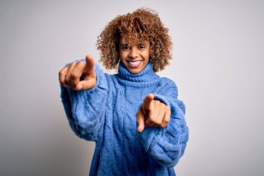 Genç ve güzel Afro-Amerikan kadın balıkçı yaka kazak giyip beyaz arka planda seni ve kamerayı işaret ediyor, pozitif ve neşeli gülümsüyor.