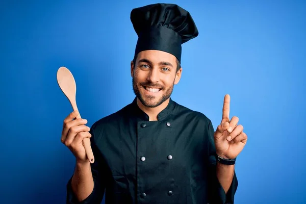 Mladý Kuchař Plnovousem Uniformě Drží Dřevěnou Lžíci Modrém Pozadí Překvapen — Stock fotografie