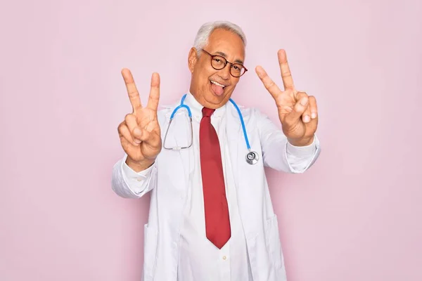 中年白发苍苍的医生 头戴听诊器 身穿专业的医疗外套 笑着伸出舌头 伸出双手的手指做胜利的标志 第二点 — 图库照片