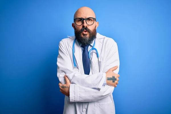 Handsome bald doctor man with beard wearing glasses and stethoscope over blue background shaking and freezing for winter cold with sad and shock expression on face