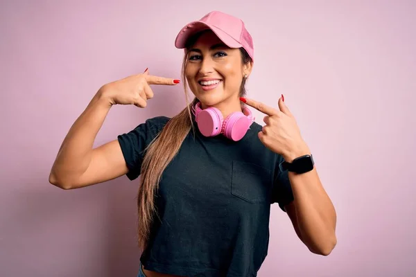 ピンクの背景にトレーニングキャップとヘッドフォンを身に着けている若い美しいフィットネススポーツの女性は 明るい表情と指の歯と口で指して笑っています 歯科衛生の概念 — ストック写真