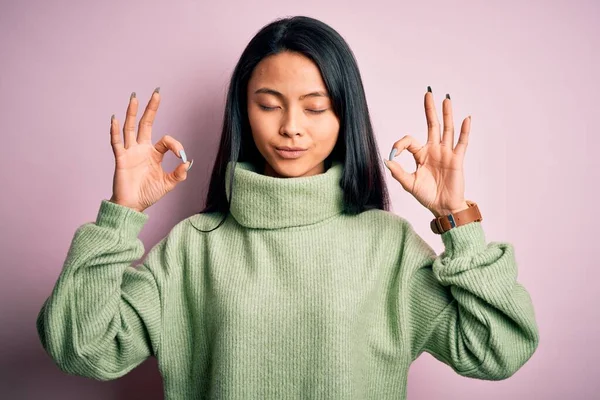孤立したピンクの背景にタートルネックセーターを身に着けている若い美しい中国の女性は 指で瞑想のジェスチャーを行う閉じて目でリラックスして笑顔 ヨガのコンセプト — ストック写真