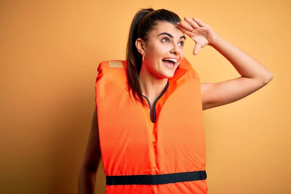 Young beautiful brunette woman wearing orange safe lifejacket over yellow background very happy and smiling looking far away with hand over head. Searching concept.