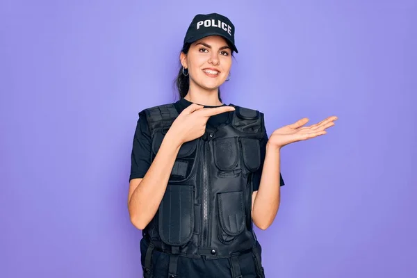 Young police woman wearing security bulletproof vest uniform over purple background amazed and smiling to the camera while presenting with hand and pointing with finger.