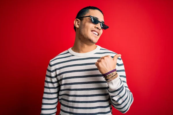 Young brazilian man wearing funny thug life sunglasses over isolated red background smiling with happy face looking and pointing to the side with thumb up.