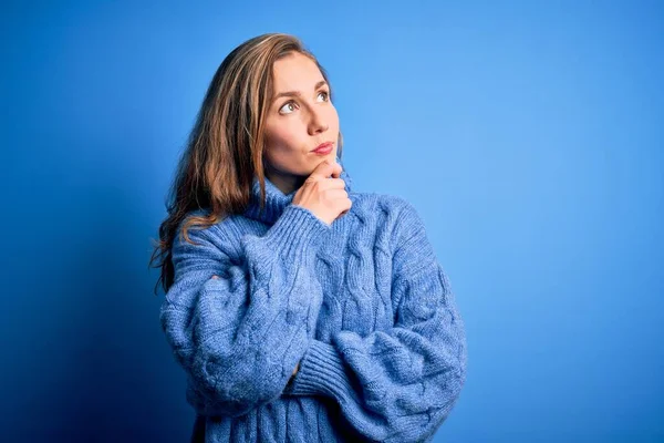 Young beautiful blonde woman wearing casual turtleneck sweater over blue background with hand on chin thinking about question, pensive expression. Smiling with thoughtful face. Doubt concept.