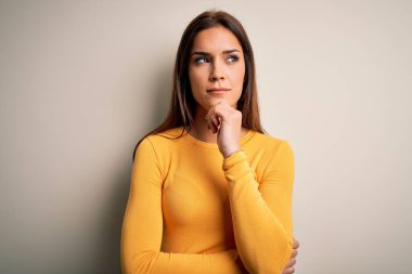 Genç, güzel esmer kadın, beyaz arka plan üzerine sarı günlük tişört giymiş, elleri çenesinde soru soran, düşünceli bir ifade takınıyor. Düşünceli bir yüzle gülümsüyor. Şüphe kavramı.