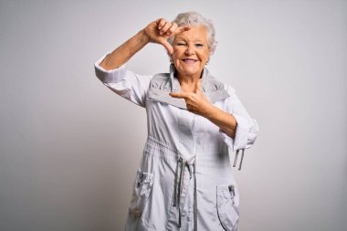 Son sınıf güzeli, gri saçlı, sıradan bir ceket giyen beyaz arka planda durup gülümseyen mutlu yüzlü elleri ve parmakları olan bir çerçeve yapan bir kadın. Yaratıcılık ve fotoğrafçılık kavramı.