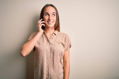 Genç, güzel bir kadın beyaz arka planda akıllı telefondan konuşuyor. Gülümsüyor ve başka tarafa bakıyor..