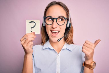Güzel çağrı merkezi ajanı kadın elinde kulaklıkla çalışıyor. Soru işareti var ve gülümseyen bir yüzle baş parmağıyla gösteriyor.