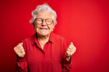 Son sınıf güzeli, gri saçlı, günlük gömlek ve gözlük takan, kırmızı arka planda mutlu ve heyecanlı bir şekilde kolları havada, gülümseyerek ve başarı için çığlık atarak kazanan bir jest yapan bir kadın. Kutlama konsepti.