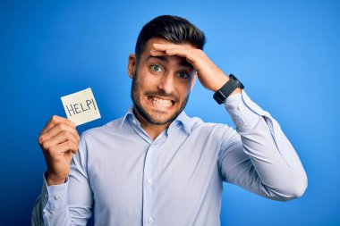 Çok çalışan genç iş adamı, mavi arka planda kağıt tutarken yardım istiyor. Elleri başının üstünde, utanç ve şaşkınlık dolu, sinirli ve hayal kırıklığına uğramış bir halde. Hata için korku ve üzüntü.