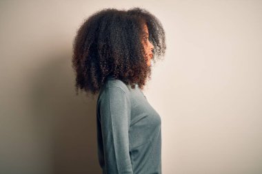 Afro saçlı genç, güzel, Afro saçlı bir kadın. Arka planda duruyor. Yan yana bakıyor, rahat bir görünüm sergiliyor..