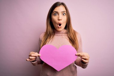 Pembe izole edilmiş arka plan üzerinde romantik, büyük kağıt kalp şeklinde bir kalp taşıyan genç sarışın kadın. Şaşırmış bir yüzle korkmuş ve korku ifadesinden heyecanlanmış.