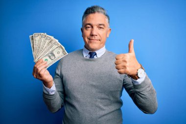 Orta yaşlı, yakışıklı, gri saçlı, elinde bir deste dolarla büyük bir gülümsemeyle mutlu, tamam işareti yapıyor, başparmağını parmaklarıyla kaldırıyor, mükemmel bir işaret.