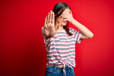 Genç ve güzel esmer kadın, günlük çizgili tişört giyiyor kırmızı arka planda duruyor, gözleri ellerle kaplıyor ve üzgün ve korku ifadeleriyle dur hareketi yapıyor. Utanç verici ve olumsuz bir kavram..