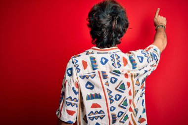 İzole edilmiş kırmızı arka plan üzerinde yaz tişörtü giyen genç ve yakışıklı bir adam geriye doğru parmak ile işaret ediyor.