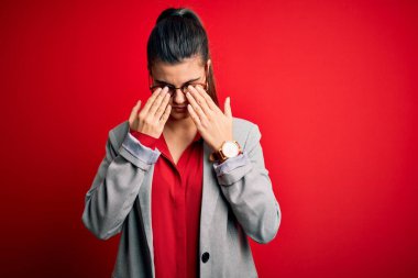 Ceket ve gözlük takan genç, güzel esmer iş kadını. Kırmızı arka planda. Yorgunluk ve baş ağrısı için gözlerini ovuşturuyor. Görüş sorunu