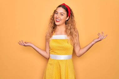 Sarı arka planda dikey duran mavi gözlü güzel sarışın kadın gülümsüyor iki elini de açık avuçlarında gösteriyor, sunuyor ve reklam karşılaştırması yapıyor ve dengede duruyor.
