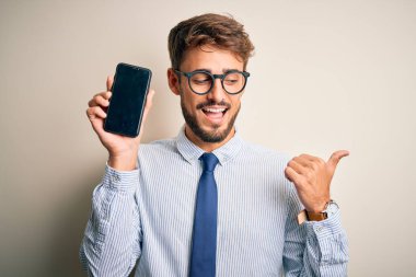 Akıllı telefonu elinde tutan genç bir işadamı izole edilmiş beyaz arka planda duruyor ve gülümseyen bir gülümsemeyle baş parmağıyla onu işaret ediyor.