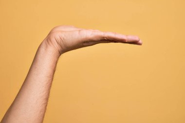 Beyaz bir gencin eli, izole edilmiş sarı arka plan üzerinde parmaklarını gösteriyor. Düz palmiye ürünü sunuyor, el hareketi yapıyor, boş kopya alanı veriyor.