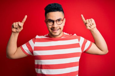 Genç ve yakışıklı bir adam, günlük çizgili tişört ve gözlük takıyor kırmızı arka planda, şaşkın, şaşkın, parmaklarıyla işaret ediyor ve kollarını kaldırıyordu..